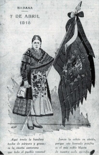 Postal de la Colonia Zamorana de Cuba en la que aparece una mujer ataviada con vestimenta tradicional y portando la Seña Bermeja, la bandera zamorana, 1918